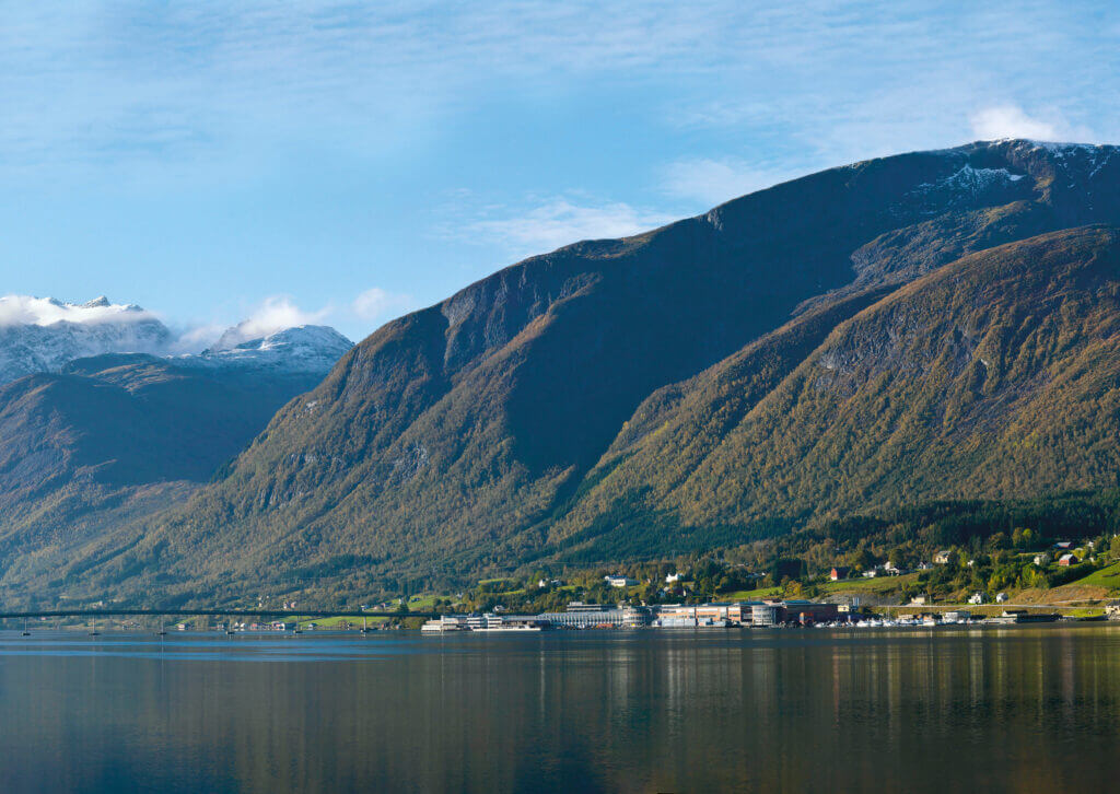 Landschaft in Norwegen, dargestellt sind Berge und ein Fjord, am Wasser liegt die Fabrik von Stressless